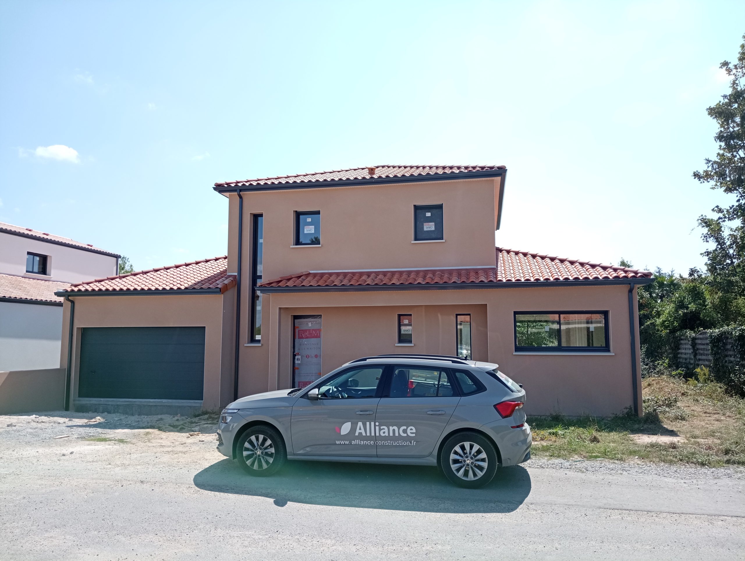 Maison moderne à étage avec crépi ocre, toit en tuiles rouges, garage intégré et voiture Alliance stationnée devant.