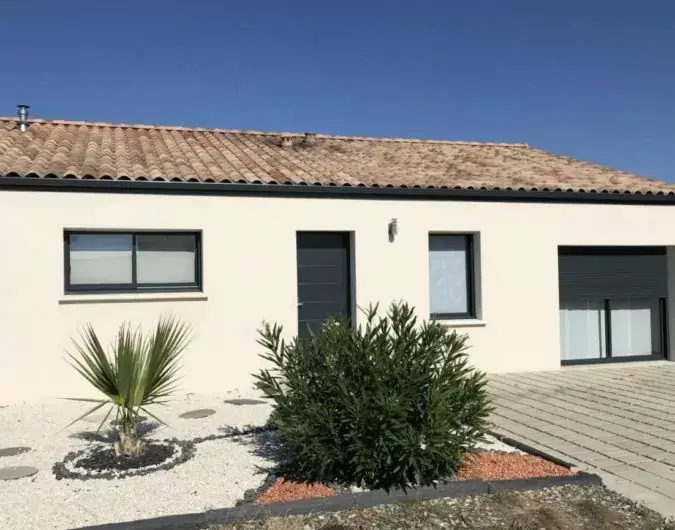 Maison de plain-pied avec toit en tuiles, façade beige et garage, agrémentée d'un jardin méditerranéen, sous un ciel bleu.