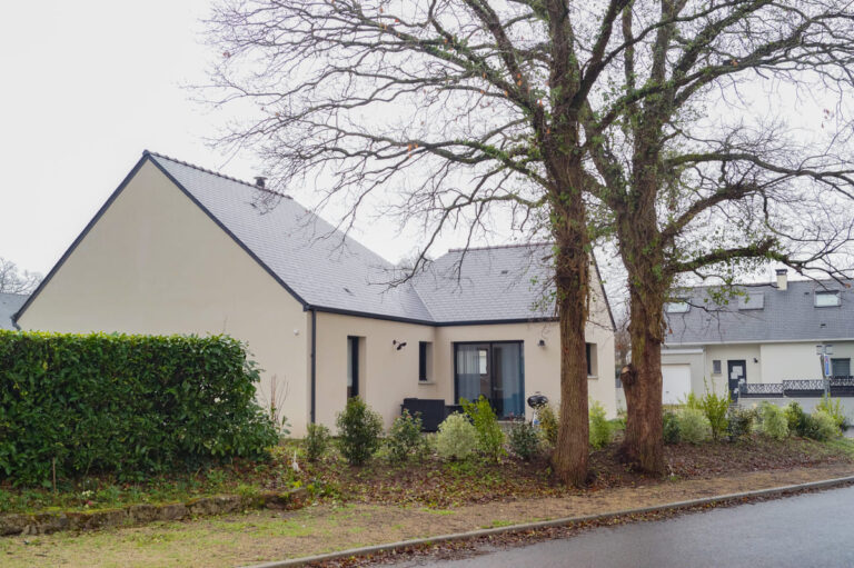 Maison de plain-pied avec toiture en ardoise, entourée de végétation et de grands arbres, sous un ciel gris.