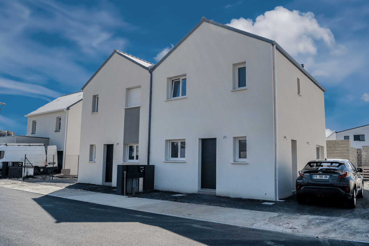 Maison mitoyenne moderne avec façade blanche, toit à deux versants et voiture stationnée devant, sous un ciel bleu lumineux.