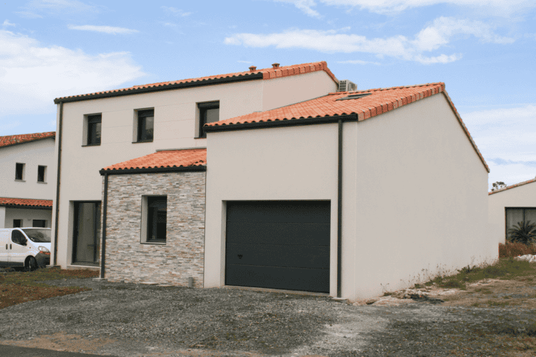 Maison contemporaine à étage avec toit en tuiles rouges, façade en pierre et garage intégré sous un ciel partiellement nuageux.
