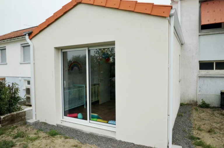 Une extension moderne avec une finition d'enduit clair, agrémentée d'une toiture inclinée en tuiles, intégrant de grandes baies vitrées pour maximiser la luminosité naturelle.