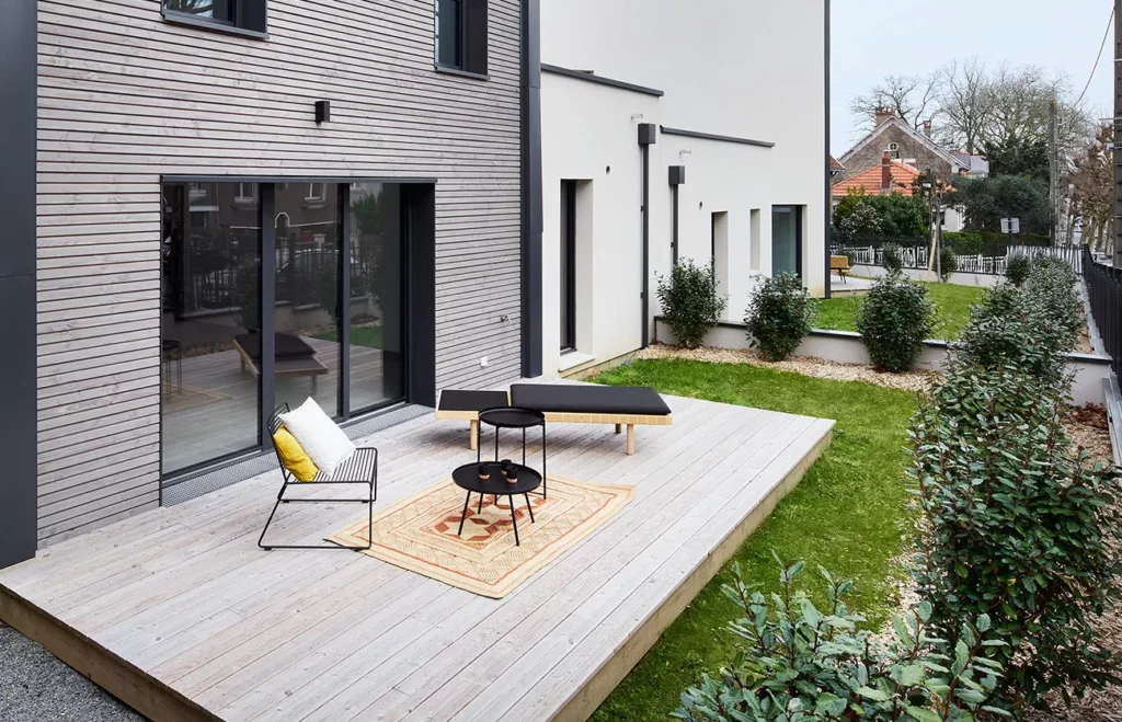 Terrasse en bois clair avec mobilier contemporain : fauteuil noir, coussin jaune, tables basses et végétation environnante.