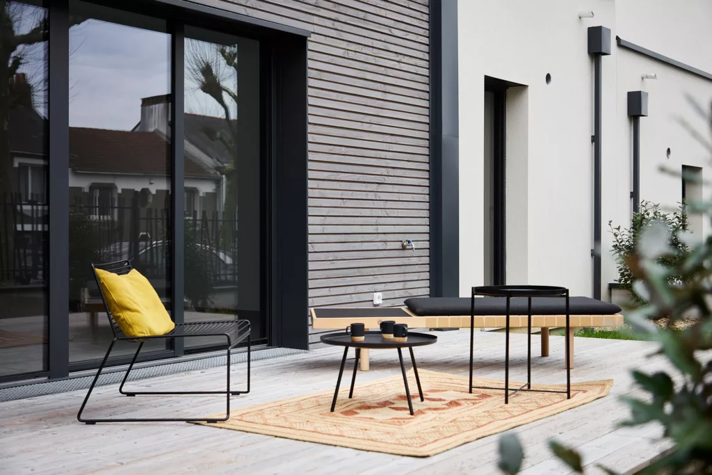 Terrasse moderne avec mobilier en métal noir, coussin jaune, table basse et tapis extérieur sur bois clair.