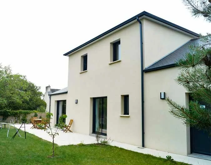 Maison contemporaine à deux étages avec toit en ardoise, façade beige, terrasse et jardin verdoyant, entourée de végétation, sous un ciel couvert.