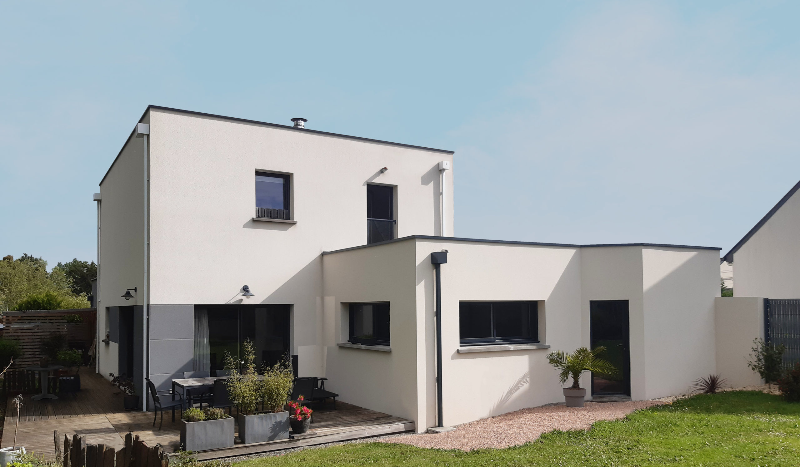 Maison moderne à toit plat avec façade en crépi blanc, grandes baies vitrées, terrasse en bois et jardin paysager.