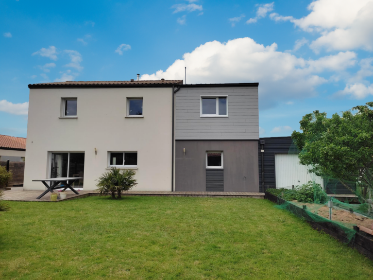 Maison rénovée avec une extension moderne, combinant une façade blanche classique et un bardage en panneaux gris, située dans un jardin arboré avec une pelouse soignée et un potager surélevé. Terrasse en bois et espace de détente aménagé pour une ambiance conviviale.