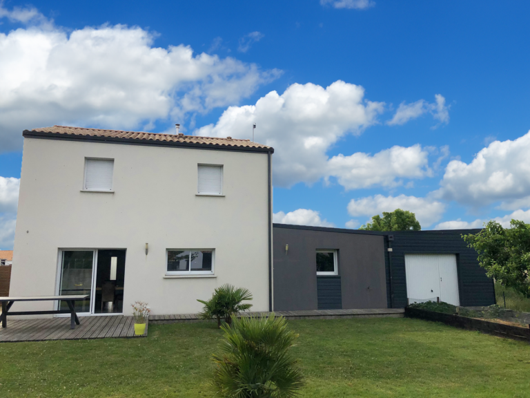 Maison avant travaux, avec une façade blanche simple et un toit en tuiles rouges. Le jardin offre une pelouse bien entretenue et une terrasse en bois, créant un espace extérieur convivial en attente d'une modernisation et d'une extension pour optimiser le style et la fonctionnalité.