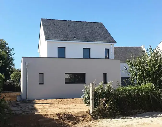 Maison traditionnelle à deux niveaux avec toiture en ardoise, façade en enduit clair et jardin en cours d’aménagement, sous un ciel bleu.