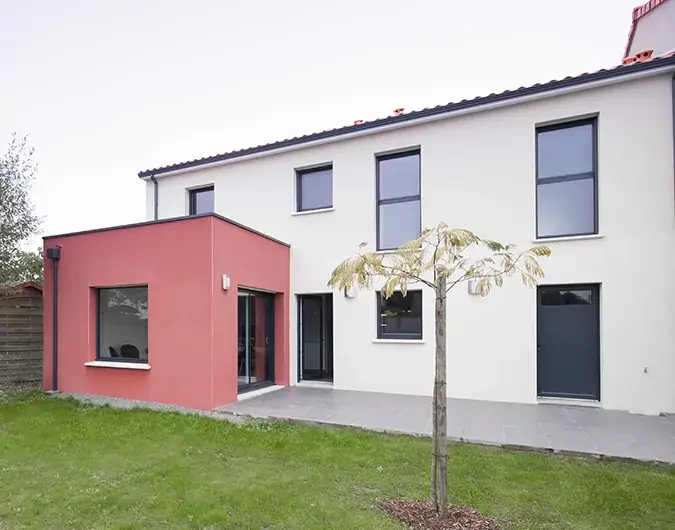 Maison à deux étages avec extension rouge, grandes fenêtres modernes et jardin verdoyant, sous un ciel couvert.