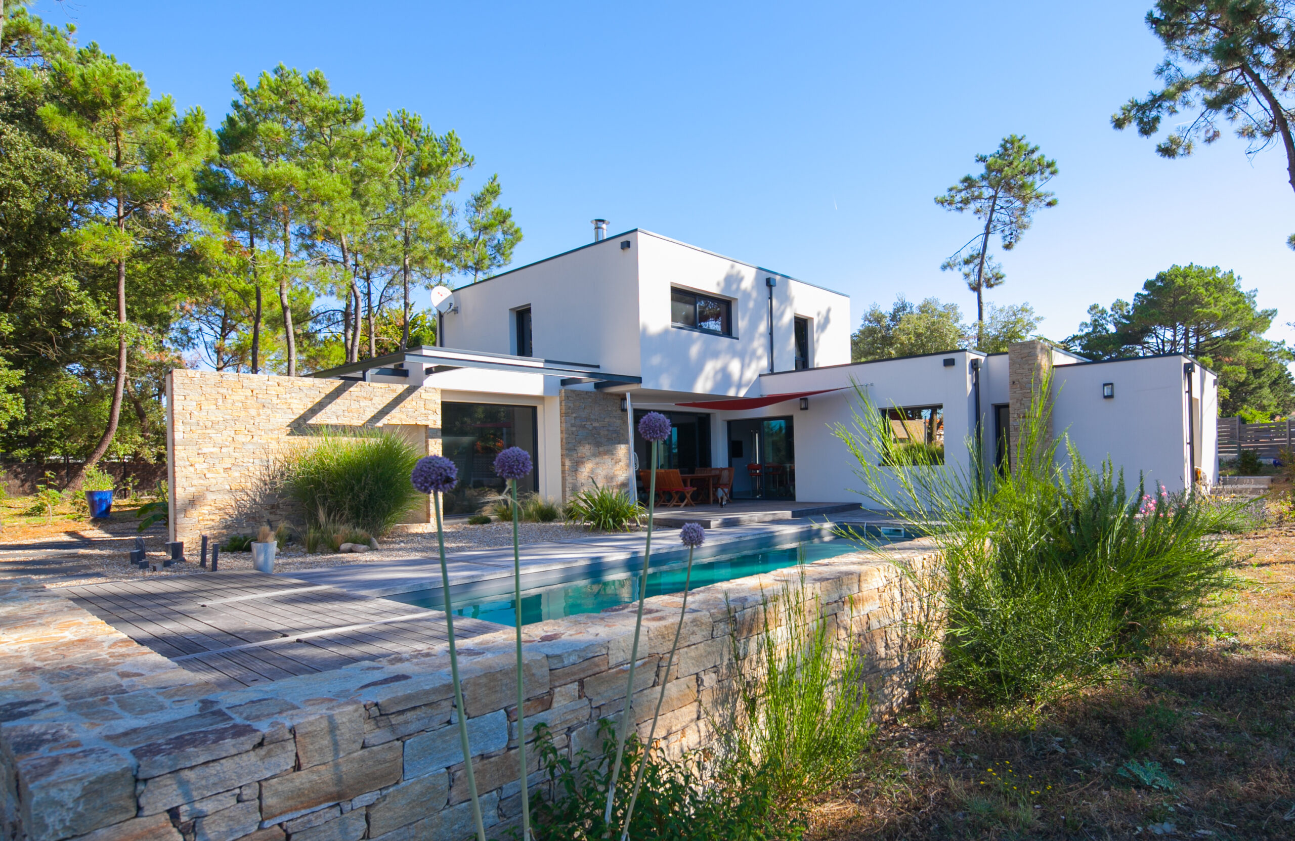 Maison contemporaine avec façade blanche, terrasse ombragée, murs en pierre naturelle, piscine élégante et jardin paysager, entourée de pins dans un cadre naturel.