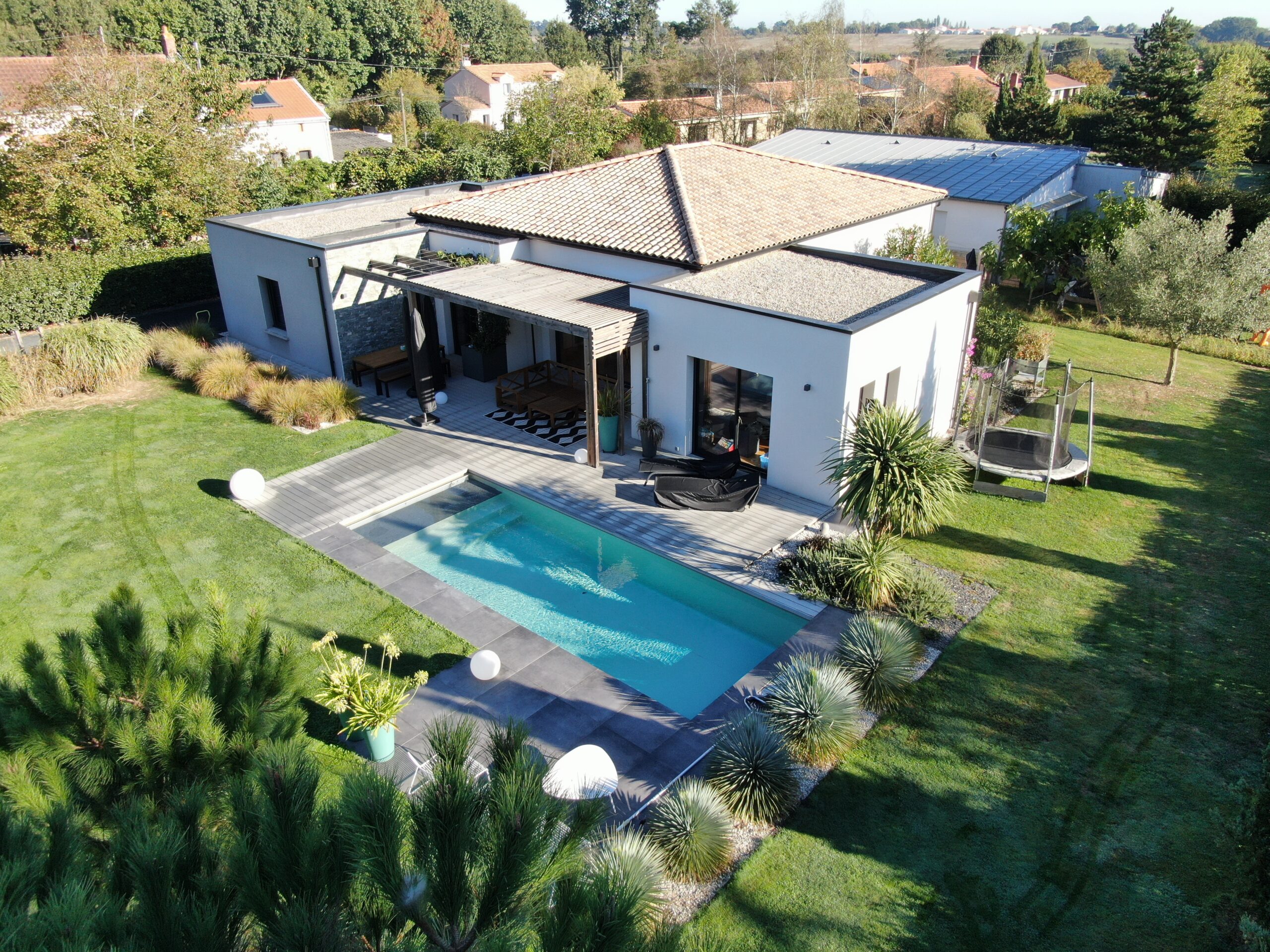 Vue aérienne d'une maison contemporaine avec toit en tuiles, terrasse aménagée, pergola en bois, piscine design entourée de végétation, et jardin paysager dans un environnement résidentiel verdoyant.