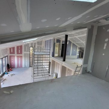 Vue d'une pièce en rénovation avec un espace ouvert. Les murs en placo sont en finition avec des bandes de joint visibles. L'échafaudage est utilisé pour le travail de finition du plafond et des murs. Les fenêtres offrent une belle lumière naturelle.