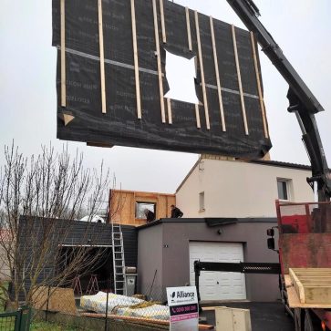 Levage d’un panneau d’ossature bois isolé à l’aide d’une grue, dans le cadre de l’extension d’une maison. Les ouvriers travaillent sur la structure en cours d'installation, avec le chantier bien organisé en arrière-plan, incluant le garage et les matériaux entreposés.