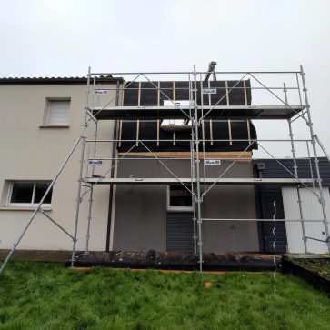 Travaux d'extension d'une maison avec un échafaudage installé devant la façade. L'ossature en bois de l'extension est visible, recouverte d'un isolant performant en préparation pour le bardage. Le chantier met en avant une transformation moderne et structurée.