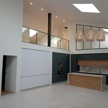 Cuisine moderne réalisée dans un espace lumineux avec un plafond haut. L'aménagement comprend des meubles de cuisine intégrés, une crédence en marbre et un îlot central en bois avec des éléments noirs. Des luminaires suspendus en osier ajoutent une touche chaleureuse. La grande baie vitrée et le puits de lumière apportent de la lumière naturelle dans cette pièce rénovée.