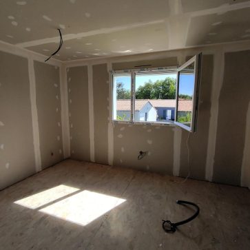 Chambre en cours de rénovation avec des murs en plaques de plâtre prêts à être peints et un sol en OSB. Une fenêtre ouverte laisse entrer la lumière naturelle, offrant une vue dégagée sur les maisons voisines. Les travaux électriques sont en préparation pour un espace moderne et fonctionnel.