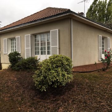 Vue de la façade avant la rénovation d'une maison unifamiliale avec un jardin bien entretenu, des fenêtres avec volets et un toit en tuiles. Le terrain est légèrement en pente, avec des plantations et des éléments paysagers visibles sur le côté. La maison, située dans un environnement calme, présente un enduit extérieur classique à rénover dans le cadre d'un projet d'amélioration de l'habitat.