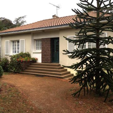 Photo avant rénovation d'une maison individuelle avec façade classique, volets blancs, escalier d'entrée en béton et jardin arboré. Projet de réhabilitation à venir pour moderniser l'extérieur.
