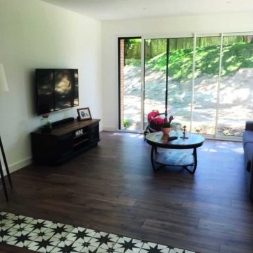 Voici le texte alternatif SEO pour l'image fournie : Salon rénové avec un sol en parquet moderne, télévision murale et baie vitrée ouvrant sur un extérieur verdoyant.