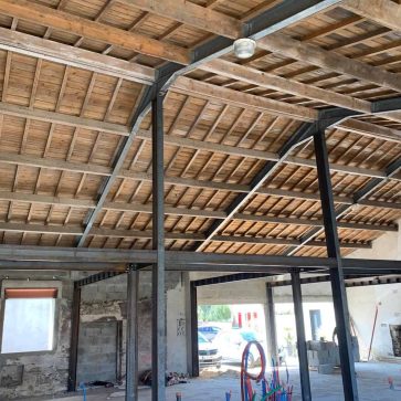 Vue intérieure d'un bâtiment en rénovation, montrant la charpente métallique et bois avec des poutres apparentes et des ouvertures en cours de finition.