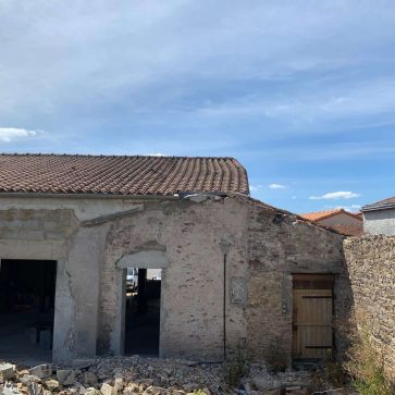 Vue extérieure d'une maison en cours de rénovation, montrant une partie de la façade détruite, avec des matériaux de construction au sol et une ancienne porte à l'arrière-plan.