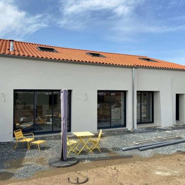 Façade d'une maison moderne avec une terrasse aménagée, comprenant des fenêtres panoramiques noires, un toit en tuiles rouges, et une unité de climatisation extérieure installée