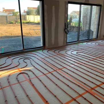 Installation de chauffage au sol avec des tuyaux posés en serpentin, prêts pour la couverture du sol, dans une pièce lumineuse avec grandes baies vitrées.