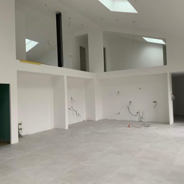 Intérieur d'un salon lumineux avec grandes baies vitrées en métal noir, sol en carrelage clair, et murs blancs dans une pièce en cours d'aménagement.