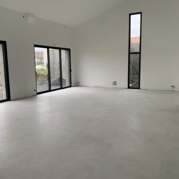 Intérieur d'un salon lumineux avec grandes baies vitrées en métal noir, sol en carrelage clair, et murs blancs dans une pièce en cours d'aménagement.