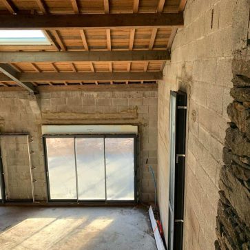 Rénovation intérieure d'un espace avec murs en parpaing, fenêtres en grande baie vitrée et toit en charpente apparente, apportant lumière naturelle à la pièce