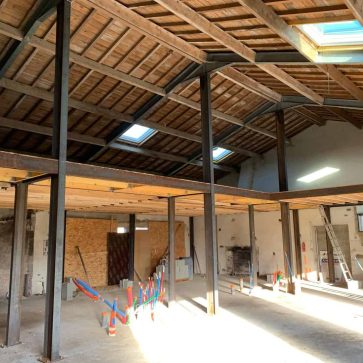 Rénovation intérieure d'un bâtiment industriel : vue sur la structure métallique avec poutres en acier, plafond en bois et verrières de toit, sol préparé pour la suite des travaux avec tuyaux et matériel en place.