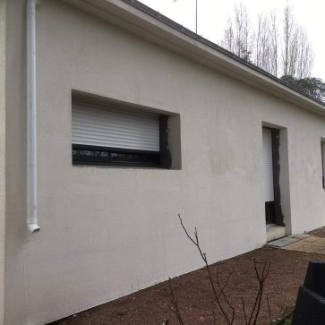 Façade d’une maison en cours de rénovation avec un enduit en phase de séchage. Les contours des fenêtres et des portes sont en attente de finition, et une descente pluviale blanche a été récemment installée. Les travaux visent à améliorer l’esthétique et l’étanchéité du bâtiment.
